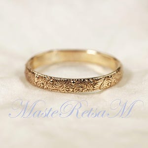 May include: A gold ring featuring an intricate, repeating engraved floral pattern along the entire outer band. The metal has a polished finish, and the design consists of delicate, swirling leaf-like motifs that wrap around the circumference.