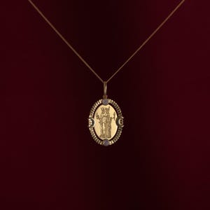 Può includere: Una collana con pendente dorato con un ciondolo ovale. Il ciondolo presenta un disegno inciso di due figure all'interno di una cornice ovale. Piccole pietre rotonde e chiare sono incastonate sul ciondolo. La collana è appesa su uno sfondo rosso scuro.