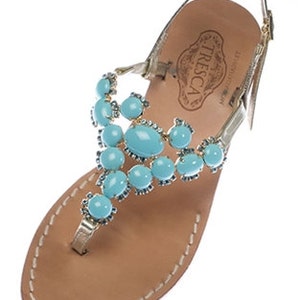 May include: Gold leather sandals with a turquoise gemstone embellishment. The sandals have a strap that goes around the ankle and a toe thong. The brand name "TRESCA" is visible on the insole.