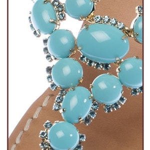 May include: Close-up of a pair of sandals featuring turquoise blue gemstone embellishments. The sandals have a leather strap and are adorned with gold-toned metal accents.