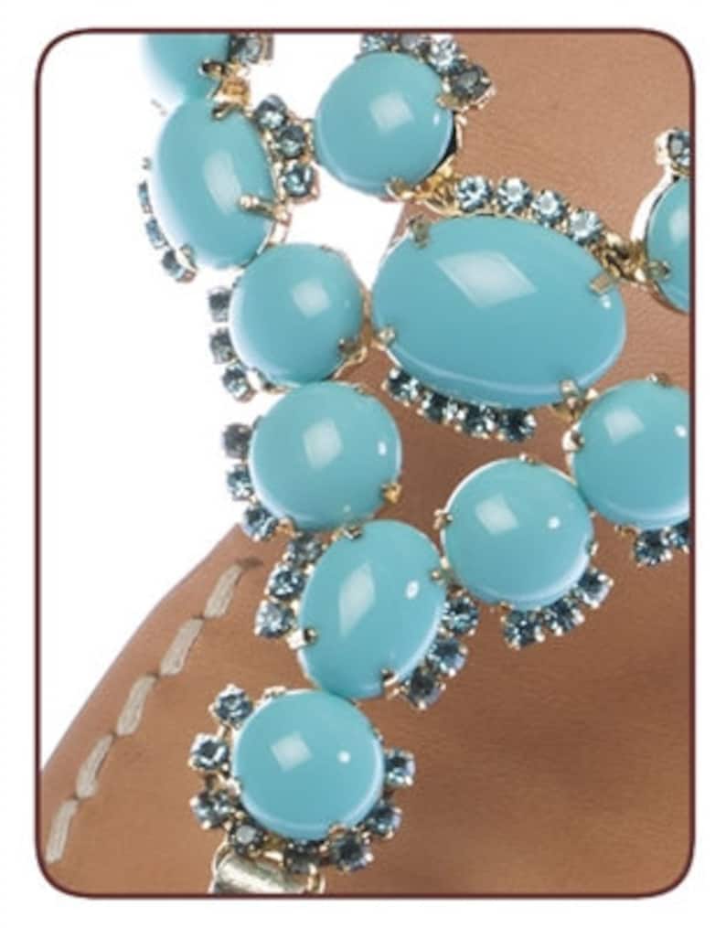 May include: Close-up of a pair of sandals featuring turquoise blue gemstone embellishments. The sandals have a leather strap and are adorned with gold-toned metal accents.