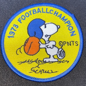 May include: A yellow and blue embroidered patch with the text "1973 Football Champion" and a cartoon of Snoopy holding a football. The text "OPNTS" is also included on the patch.