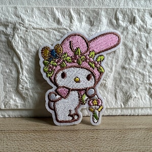 Puede incluir: Parche bordado con un personaje de dibujos animados con una capucha rosa con forma de conejo y detalles florales. El personaje es blanco con contornos marrones y sostiene un pequeño ramo. Fondo blanco.