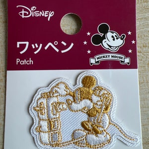 Può includere: Una toppa da stirare bianca ricamata con Mickey Mouse in filo dorato. La toppa è su una carta bianca con il testo "Disney" e "Mickey Mouse" in nero. Il testo "Patch" e "フッペン" è anche sulla carta.
