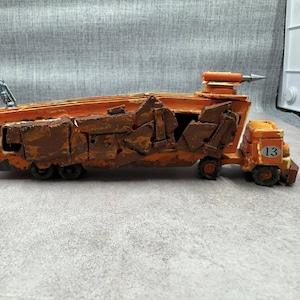May include: A handmade, orange toy truck with brown, textured details. The truck has a number 13 on the side and a silver crane arm. The truck is a unique, artistic creation.