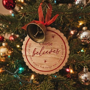 May include: A round wooden Christmas ornament with a scalloped edge, featuring a metal bell attached by a red ribbon. The ornament is engraved with the name Charlie, the word believes, and the text The bell still rings for those who truly believe.