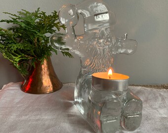 May include: A clear glass Santa Claus candle holder with a candle inside. The Santa Claus is holding a bell in his left hand and has a beard and a hat. The candle holder is made by Boda Nova.
