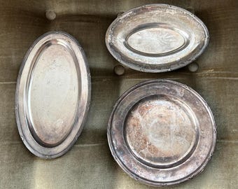 Può includere: Tre piatti da portata d'argento di diverse dimensioni e forme. I piatti sono ovali e rotondi, con un bordo rialzato e una superficie liscia e riflettente.