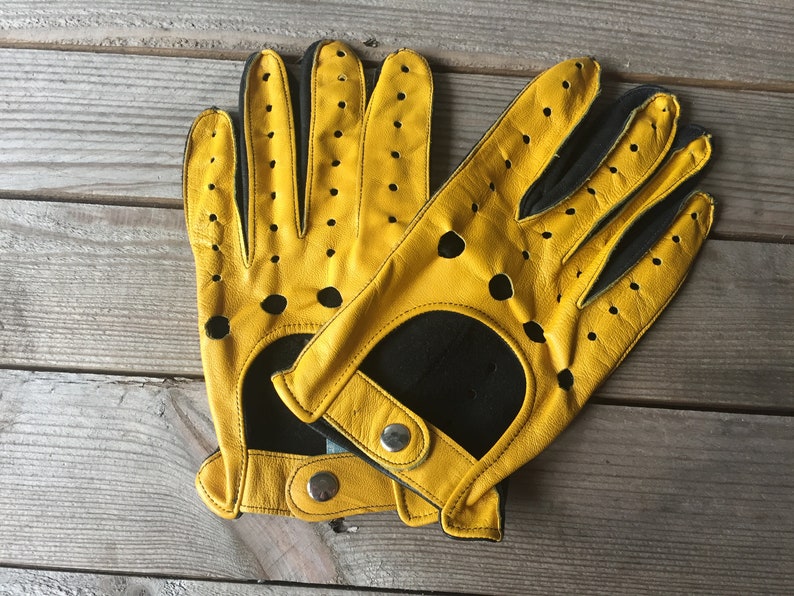 May include: A pair of yellow leather driving gloves with black accents and perforations. The gloves have a snap closure on the wrist.