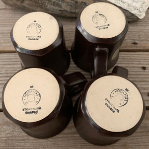 Puede incluir: Cuatro tazas de gres marr&oacute;n con un esmalte blanco y un logotipo negro que dice "Hogan&auml;s Stoneware Sweden".