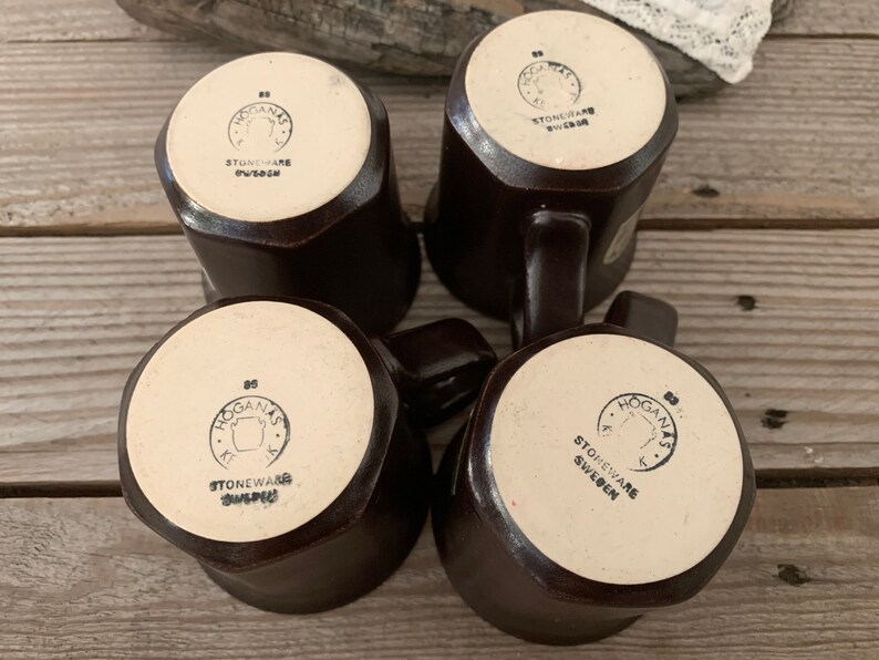 Puede incluir: Cuatro tazas de gres marr&oacute;n con un esmalte blanco y un logotipo negro que dice "Hogan&auml;s Stoneware Sweden".