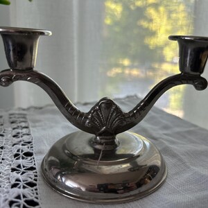 May include: A silver-colored, two-arm candlestick holder. The base is round, supporting two curved arms that hold the candle cups. The design includes a decorative, shell-like element at the center. The candlestick holder is made of metal.