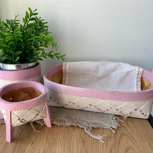 May include: A set of decorative birch bark baskets and a small potted plant. The baskets are painted white with pink trim and have a woven design. The plant is in a metal pot with a pink and white painted exterior.