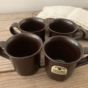 Puede incluir: Cuatro tazas de cer&aacute;mica marr&oacute;n con asas. Las tazas son todas del mismo tama&ntilde;o y forma. Las tazas tienen un acabado liso y mate. Las tazas est&aacute;n dispuestas en forma de cuadrado. Las tazas est&aacute;n sobre una superficie de madera. El texto en la taza dice "Hogan&auml;s,  Byr&aring;n engosta,  Utm&auml;rkt maskindiskbart,  H&ouml;ganschina,  Sverige".