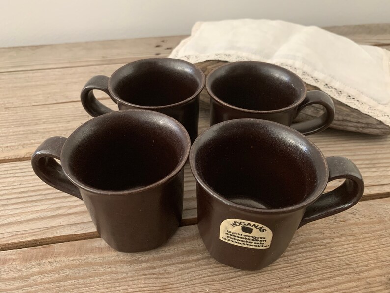 Puede incluir: Cuatro tazas de cer&aacute;mica marr&oacute;n con asas. Las tazas son todas del mismo tama&ntilde;o y forma. Las tazas tienen un acabado liso y mate. Las tazas est&aacute;n dispuestas en forma de cuadrado. Las tazas est&aacute;n sobre una superficie de madera. El texto en la taza dice "Hogan&auml;s,  Byr&aring;n engosta,  Utm&auml;rkt maskindiskbart,  H&ouml;ganschina,  Sverige".