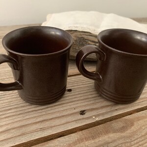 May include: Two brown ceramic mugs with handles. The mugs are on a wooden surface.
