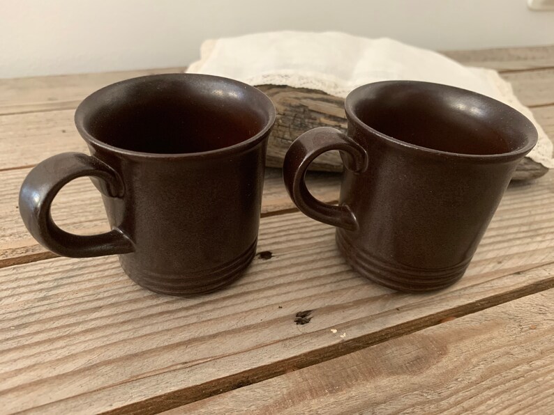May include: Two brown ceramic mugs with handles. The mugs are on a wooden surface.