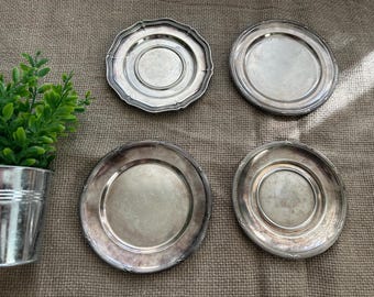 Può includere: Quattro sottobicchieri antichi in argento con bordi decorativi sono disposti su una superficie marrone strutturata. Un sottobicchiere ha un bordo smerlato, mentre gli altri hanno bordi arrotondati. Una piccola pianta in vaso è visibile a sinistra.