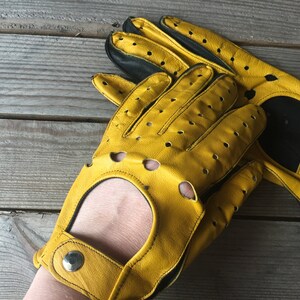 May include: A pair of yellow leather driving gloves with black accents and perforations. The gloves have a wrist strap with a snap closure.