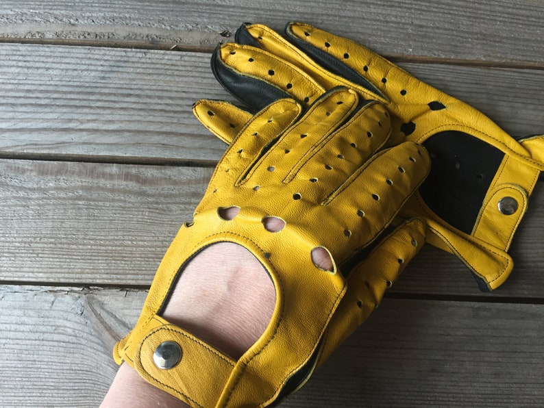 May include: A pair of yellow leather driving gloves with black accents and perforations. The gloves have a wrist strap with a snap closure.