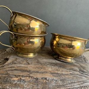May include: Three brass cups with handles. The cups are stacked on top of each other. The cups are shiny and reflect light.