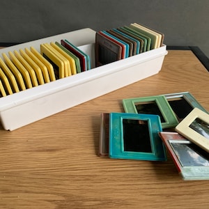 Peut inclure: Une boîte de rangement blanche en plastique pour diapositives avec une variété de diapositives colorées à l'intérieur. La boîte est posée sur une surface en bois avec plusieurs diapositives éparpillées autour. Les diapositives sont de différentes couleurs, notamment le vert, le bleu et le jaune.