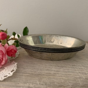 May include: A set of four silver pie dishes stacked on top of each other. The dishes are round and have a slightly scalloped edge. They are sitting on a light wood surface.
