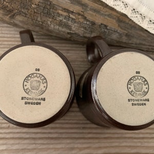 May include: Two brown stoneware mugs with white bottoms. The bottoms of the mugs have a brown circle with the text "Hogan&auml;s Keramik" and the words "Stoneware Sweden".
