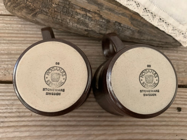 May include: Two brown stoneware mugs with white bottoms. The bottoms of the mugs have a brown circle with the text "Hogan&auml;s Keramik" and the words "Stoneware Sweden".