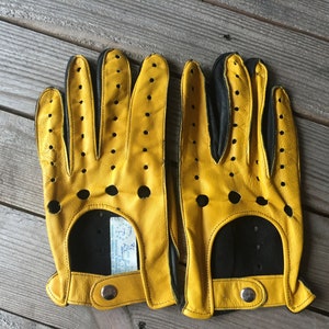May include: A pair of yellow leather driving gloves with black accents and perforations. The gloves have a snap closure on the wrist.