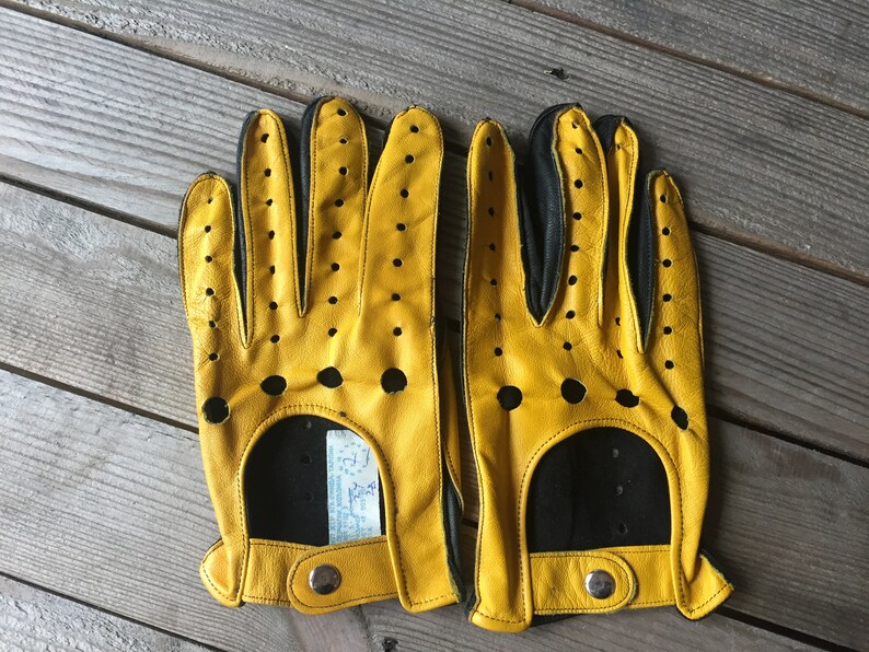 May include: A pair of yellow leather driving gloves with black accents and perforations. The gloves have a snap closure on the wrist.