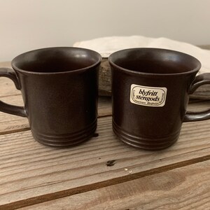 May include: Two brown ceramic mugs with a white label on one mug that reads "blyfritt stengods".