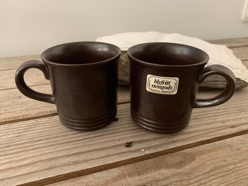 May include: Two brown ceramic mugs with a white label on one mug that reads "blyfritt stengods".