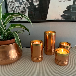 May include: A collection of copper candle holders with lit candles, featuring a gold rim. The holders vary in size and are arranged on a cream-colored surface. A copper planter with a green and white plant is also visible.