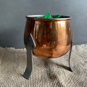 May include: A copper pot with a shiny finish sits on a metal stand with three legs. The pot is empty and has a small green plant inside.