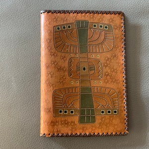 May include: A brown leather journal with a geometric design in gold and green. The design features a central vertical line with a series of squares and circles.