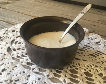 May include: A dark brown ceramic sugar bowl filled with white granulated sugar, with a silver spoon resting inside. The bowl sits on a white, open-weave doily, with a wooden surface in the background.