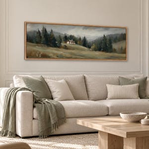 May include: A panoramic landscape painting in a light wood frame depicting a small house nestled in a grassy field surrounded by dark green pine trees under a cloudy, overcast sky, mounted on a wall above a beige sofa with green pillows.