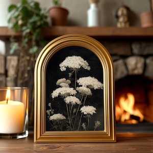 May include: A framed artwork featuring white Queen Anne's Lace flowers against a black background. The gold-colored frame has an arched top. A lit candle in a glass holder is to the left of the frame. The background includes a fireplace and a wooden shelf.