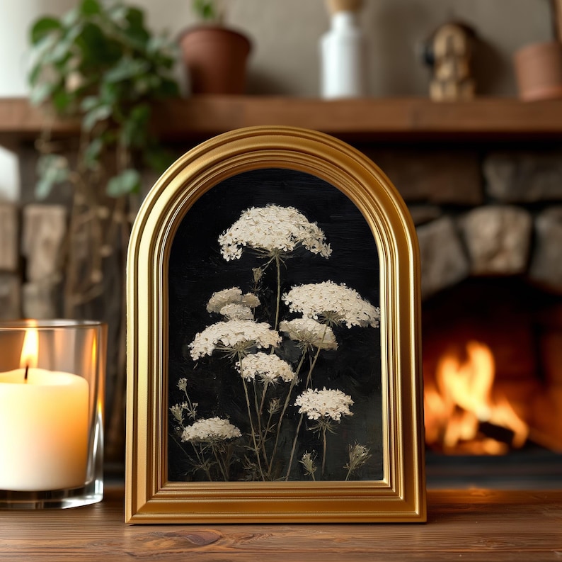 May include: A framed artwork featuring white Queen Anne's Lace flowers against a black background. The gold-colored frame has an arched top. A lit candle in a glass holder is to the left of the frame. The background includes a fireplace and a wooden shelf.