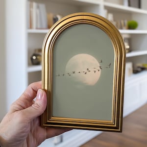 May include: An arched gold picture frame containing a print of a large, pale moon set against a muted sage green sky. A flock of dark birds is depicted flying in a horizontal formation across the center of the moon and the surrounding sky.