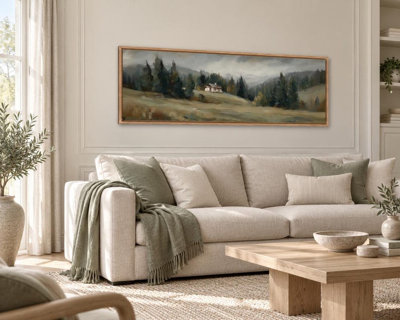 May include: A panoramic landscape painting in a light wood frame depicting a small house nestled in a grassy field surrounded by dark green pine trees under a cloudy, overcast sky, mounted on a wall above a beige sofa with green pillows.