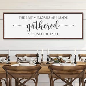 May include: A rectangular wall sign with a dark brown frame and a white background. The text reads THE BEST MEMORIES ARE MADE gathered AROUND THE TABLE in black lettering, with the word gathered written in a large, elegant cursive script font.