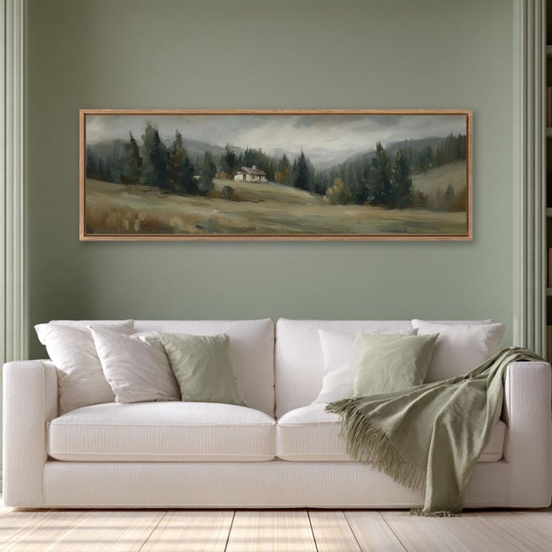 May include: A panoramic landscape painting in a wooden frame featuring a small cabin nestled in a grassy field surrounded by dark green pine trees under a cloudy, overcast sky. The artwork displays muted earth tones and soft, painterly brushstrokes.