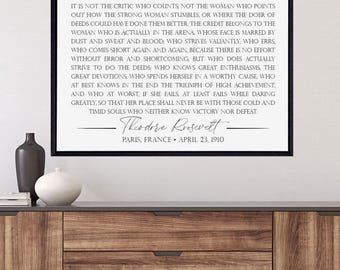 Op de afbeelding: Een zwart-witte printbare quote van Theodore Roosevelt over de vrouw in de arena. De quote luidt: "Het is niet de criticus die telt, niet de vrouw die erop wijst hoe de sterke vrouw struikelt..." De quote is ingelijst in een zwart frame en hangt aan een muur.