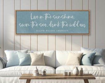 May include: A rectangular wall sign with a light blue background and a wooden frame. The text reads: Live in the sunshine, swim the sea, drink the wild air, Ralph Waldo Emerson. The lettering is in a white, cursive script font.