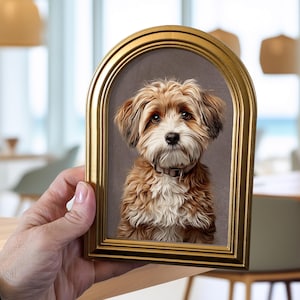 May include: An arched gold picture frame containing a portrait of a fluffy, light brown and white dog with dark eyes and a small collar. The frame has a smooth, metallic finish and a rounded top, showcasing the detailed illustration of the canine.
