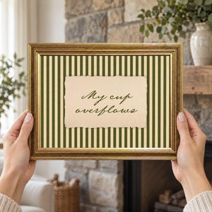 May include: A rectangular gold-toned picture frame containing a background with vertical dark green and cream stripes. Centered in the frame is a cream-colored paper insert with deckled edges featuring the black cursive text My cup overflows.