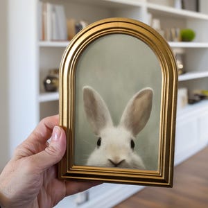 May include: A framed print featuring a white rabbit with long ears. The print is set within a gold-colored, arched frame. The rabbit's face is centered, with its eyes and nose visible. The background is a soft, muted green.