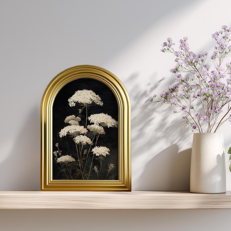 May include: A framed botanical print featuring white Queen Anne's Lace flowers against a black background. The gold-colored frame has an arched top. A vase of purple flowers sits to the right on a wooden shelf.